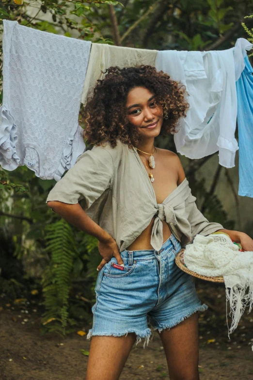 a woman standing next to a pile of clothes, by Samuel Scott, pexels contest winner, renaissance, croptop and shorts, brown curly hair, wearing a linen shirt, tropical style