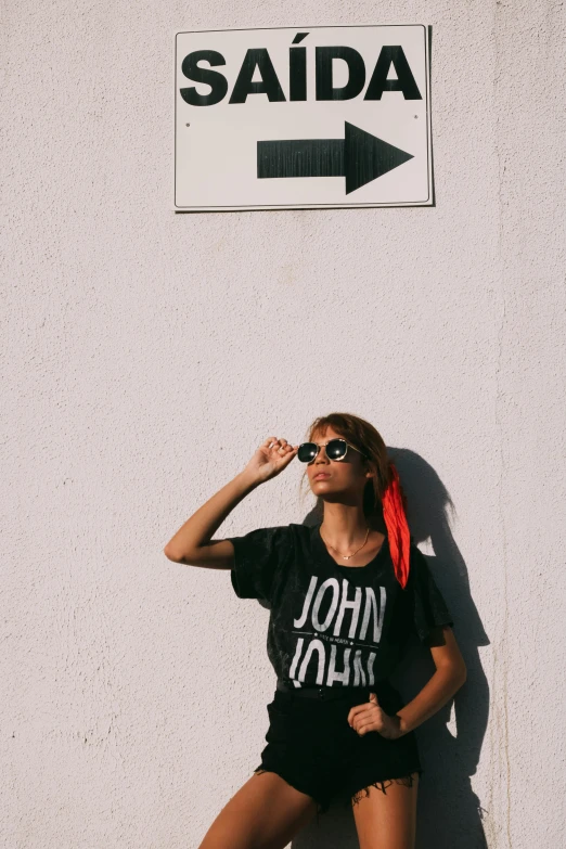 a woman leaning against a wall in front of a sign, by Julia Pishtar, trending on unsplash, graphic tees, elton john lennon, black sunglasses, filmed in the style of john toll