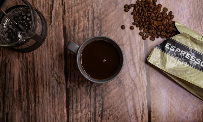 a bag of coffee next to a cup of coffee, a still life, inspired by Elsa Bleda, trending on unsplash, background image, vray 8k, on a wooden desk, render unreal engine-h 704