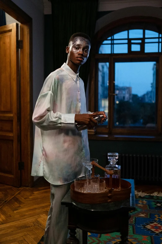 a man standing in a living room next to a table, an album cover, inspired by Barthélemy Menn, trending on unsplash, hyperrealism, wearing silver silk robe, irridescent ghostly, potions, model posing