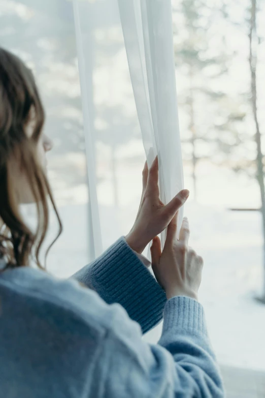 a woman in a blue sweater looking out a window, soft volume absorbation, grey, transparent, lower quality