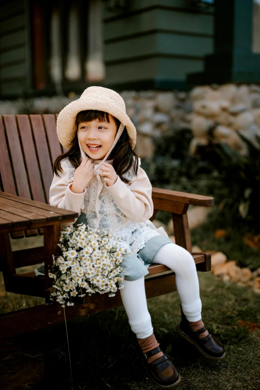 a little girl sitting on top of a wooden bench, holding a flower, in style of lam manh, smiling playfully, wearing a cute hat