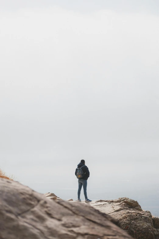 a person standing on top of a large rock, by Ryan Pancoast, unsplash, overcast gray skies, gazing off into the horizon, walking away, unfocused