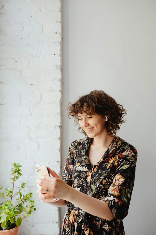 a woman standing in front of a window looking at her phone, a picture, happening, dark short curly hair smiling, with flowers and plants, 2019 trending photo, on a white table