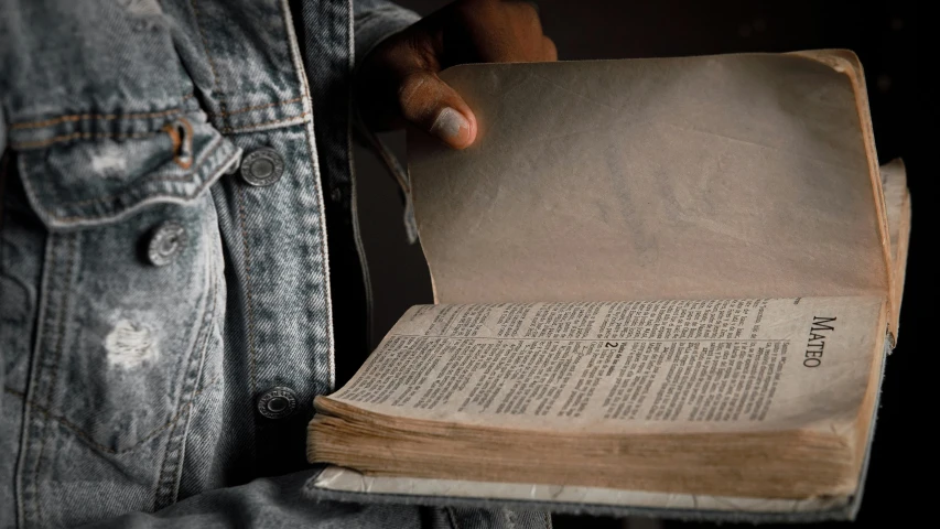 a close up of a person holding an open book, trending on unsplash, renaissance, wearing torn clothes, bible, grey, denim