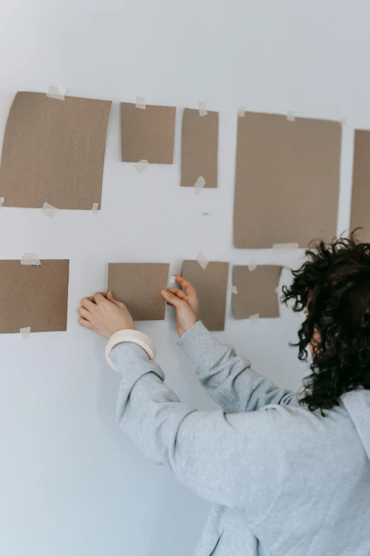 a woman standing in front of a wall covered in pieces of cardboard, a picture, trending on unsplash, square shapes, mat drawing paper, inspect in inventory image, light - brown wall