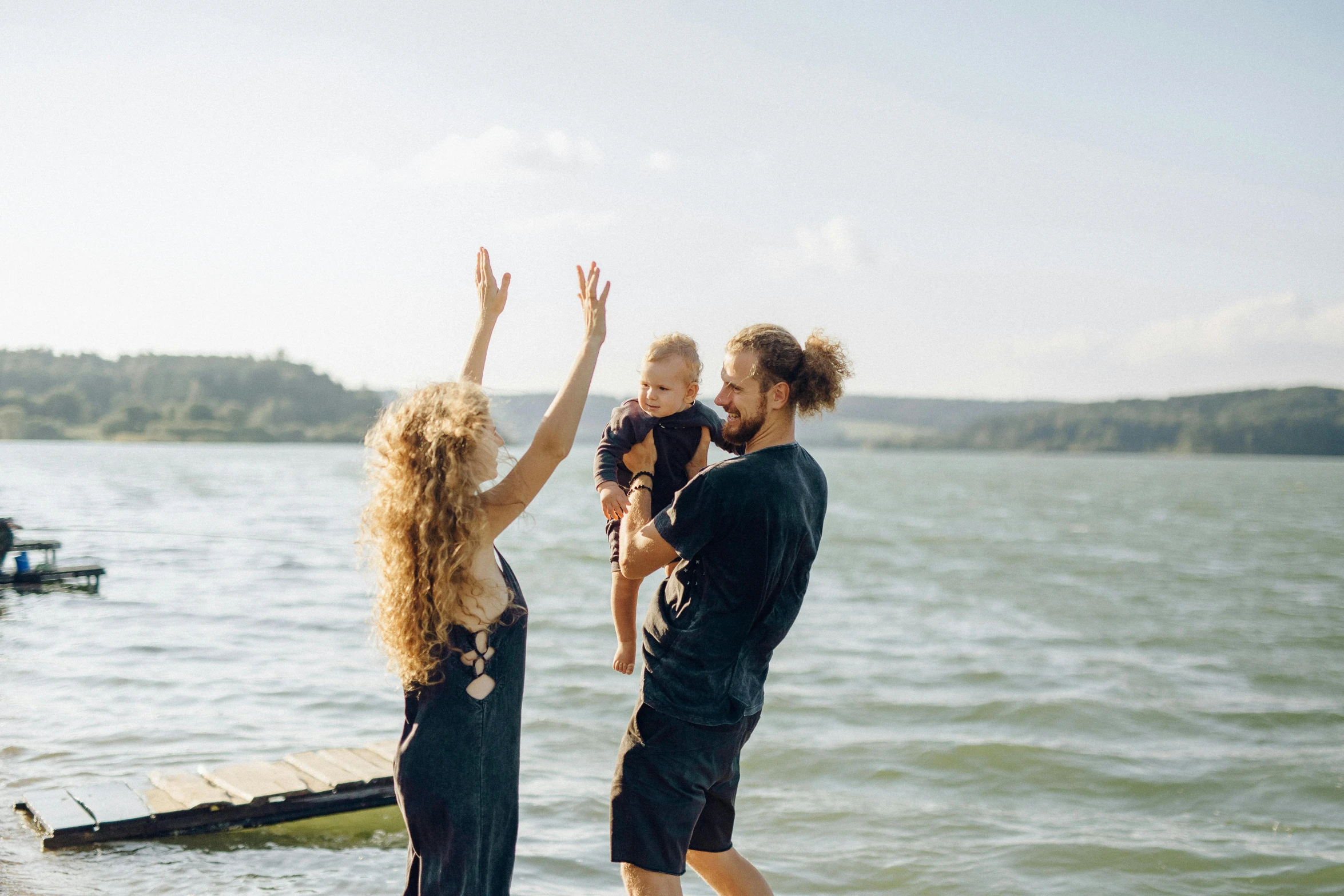 a couple of people that are standing in the water, with a kid, hands in air, profile image, view from the lake