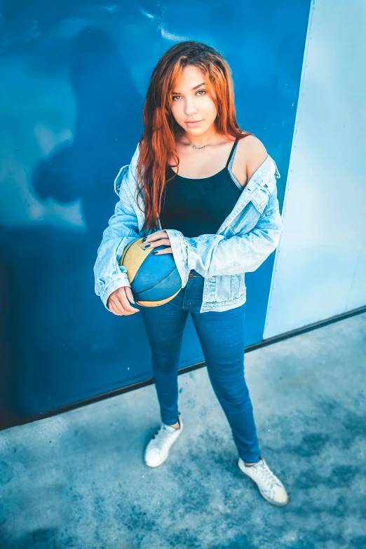 a woman standing in front of a blue wall holding a basketball, an album cover, trending on pexels, red haired girl, 15081959 21121991 01012000 4k, wearing a jeans jackets, a high angle shot