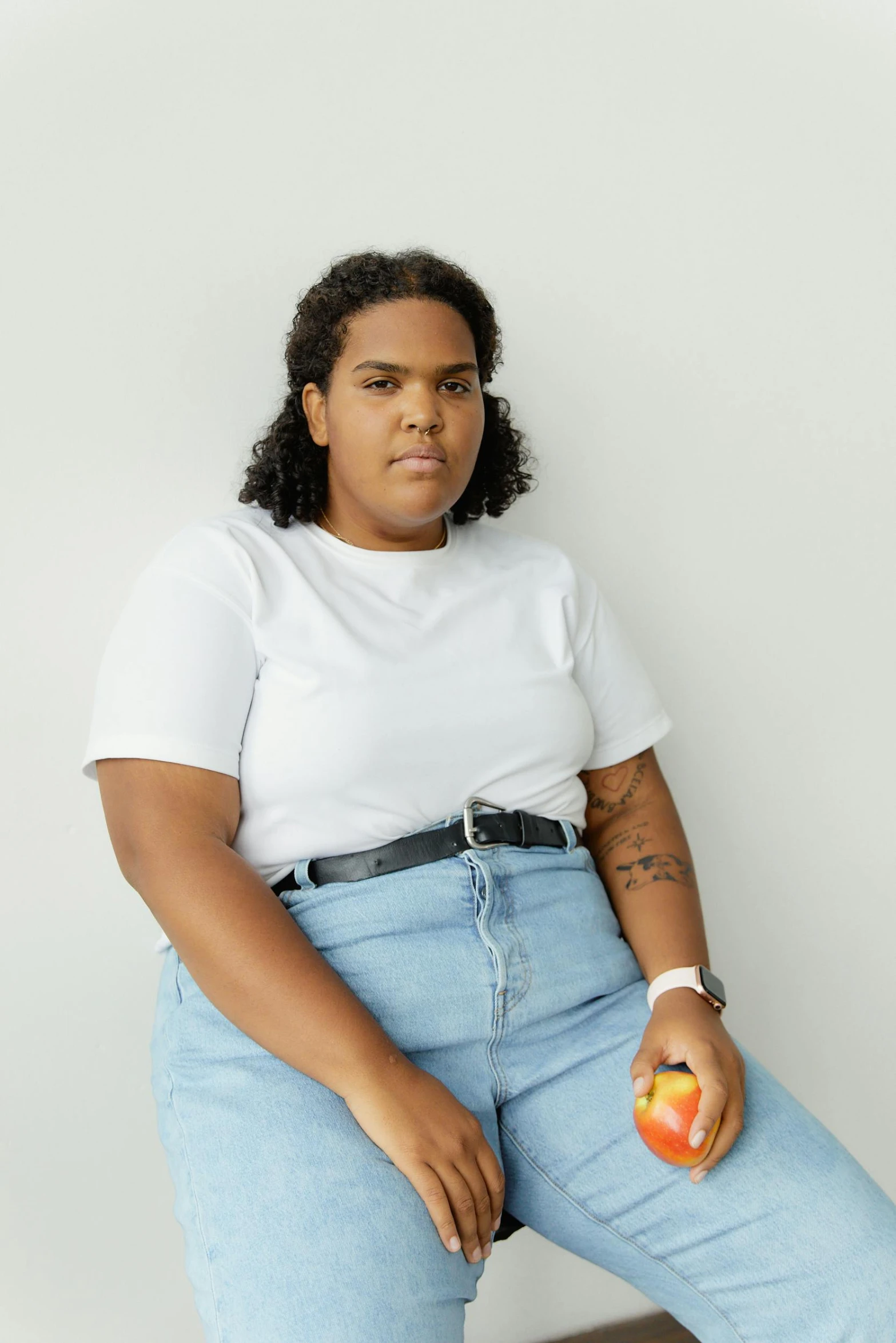 a woman sitting on a chair holding an apple, by Grace Polit, trending on pexels, tan skin a tee shirt and shorts, plus-sized, non-binary, crossed arms