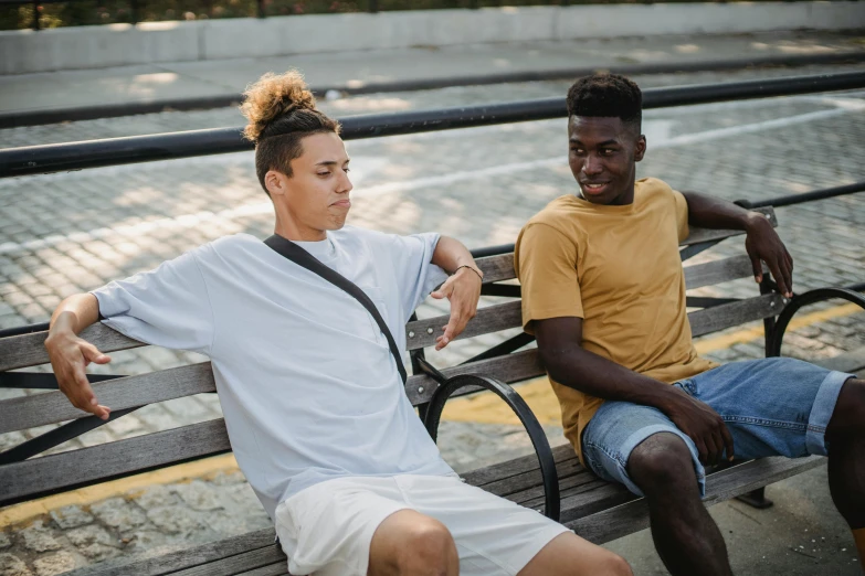 two men sitting on a bench next to each other, trending on pexels, happening, tan skin a tee shirt and shorts, black teenage boy, androgynous male, coloured