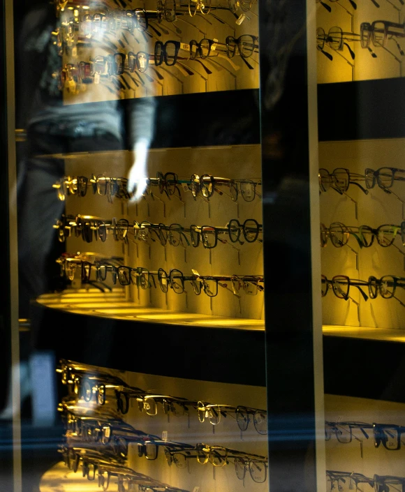 a man standing in front of a display of glasses, by Carey Morris, unsplash, gold and black color scheme, display case, yellow pupils, back lit