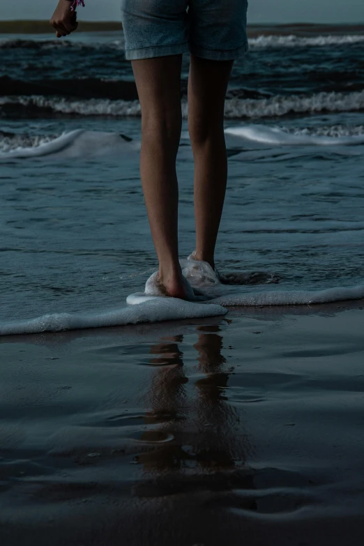 a woman standing on top of a beach next to the ocean, a picture, by Elsa Bleda, unsplash contest winner, detailed legs, foam, late night, teenage girl