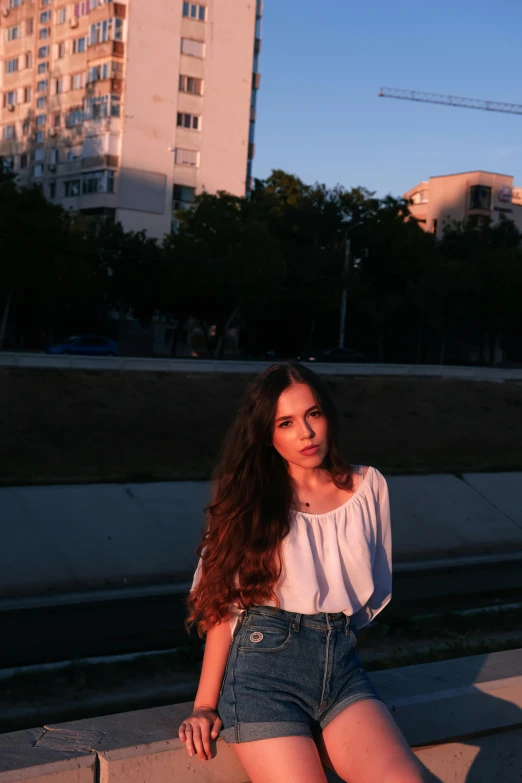 a woman sitting on a ledge in front of a building, ((sunset)), with long dark hair, estefania villegas burgos, posing for a picture