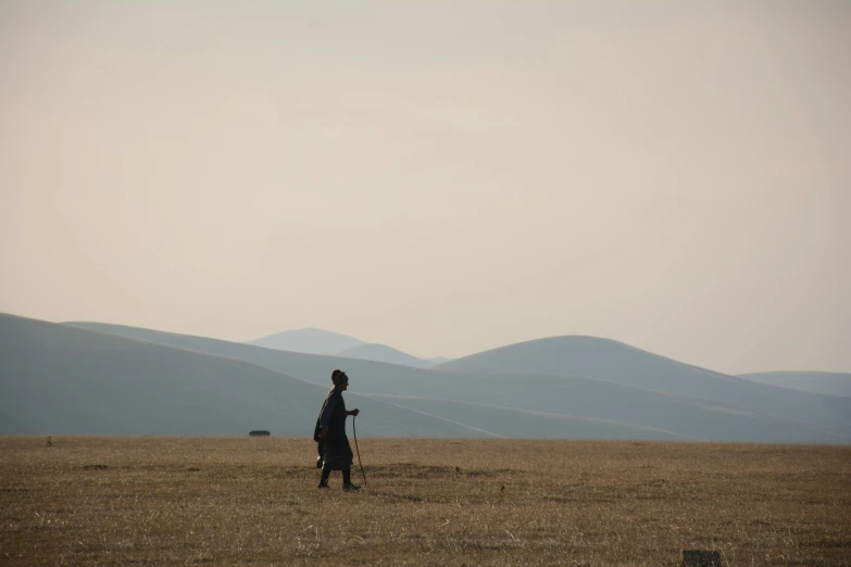 a man walking across a dry grass covered field, by Attila Meszlenyi, pexels contest winner, land art, genghis khan, sweeping vista, lone silhouette in the distance, jin shan