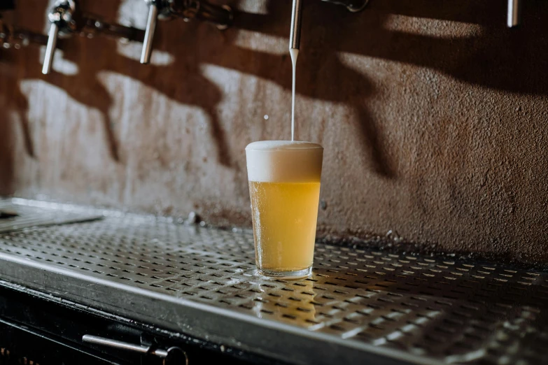 a glass of beer sitting on top of a metal counter, unsplash, private press, white and yellow scheme, shepherd's crook, pouring, inside a bar