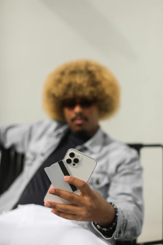 a man sitting in a chair holding a cell phone, an album cover, trending on pexels, short blonde afro, taken on iphone 14 pro, long afro hair, recognizable