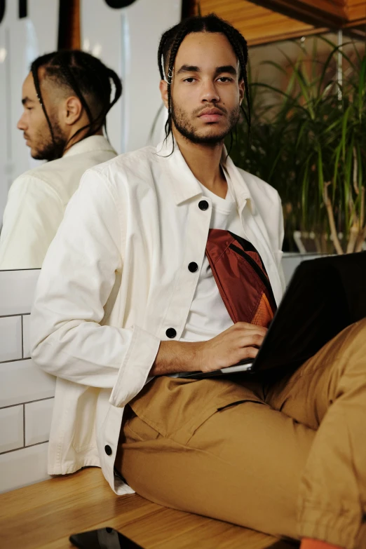 a man sitting on a table with a laptop, an album cover, trending on pexels, light brown trenchcoat, lab coat and tee shirt, holding notebook, masculine appeal high fashion