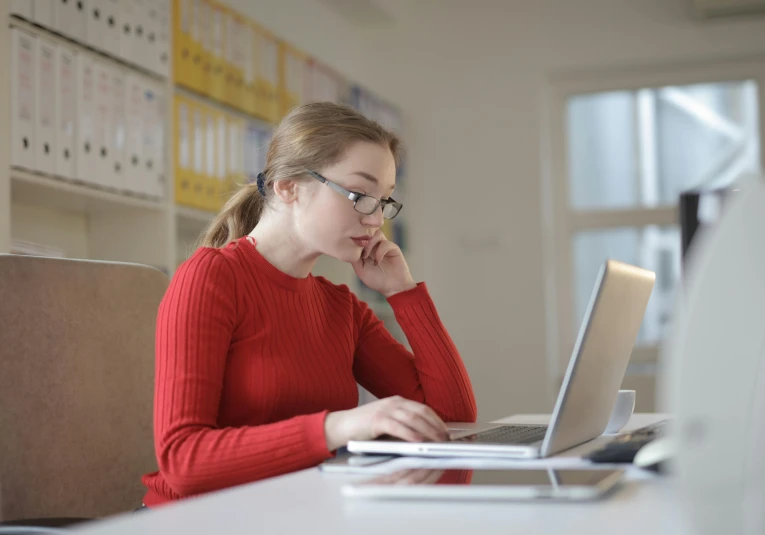 a woman sitting at a desk using a laptop computer, pexels, wearing red formal attire, girl wearing round glasses, thumbnail, studious