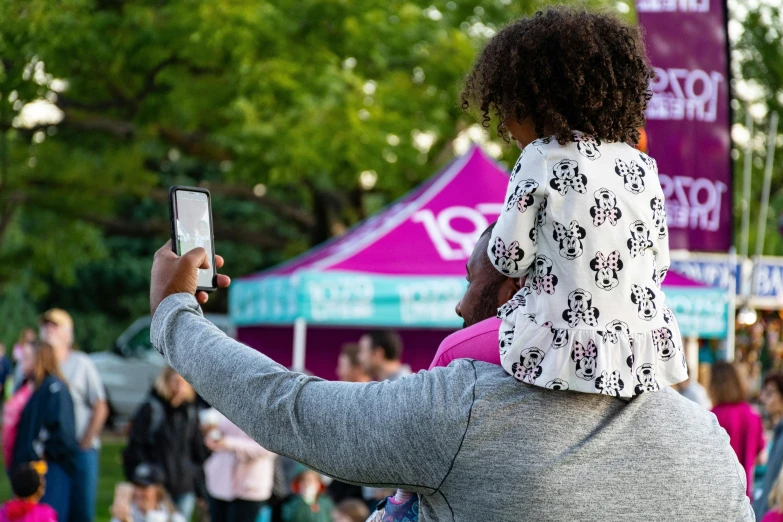 a woman holding a child up to take a picture, looking at his phone, community celebration, square, performing