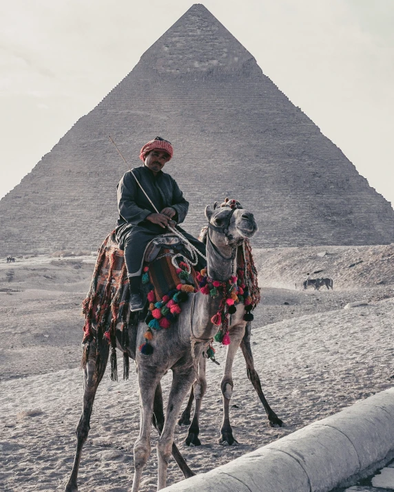 a man riding on the back of a camel in front of a pyramid, pexels contest winner, aida muluneh, ornately dressed, winter, looking serious