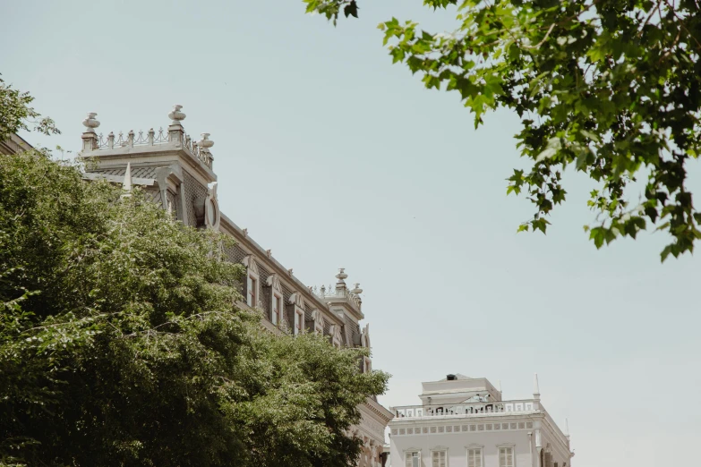 a group of people walking down a street next to a tall building, a photo, by Pablo Rey, pexels contest winner, neoclassicism, branches and foliage, summer sky, background image, ornate palace made of green