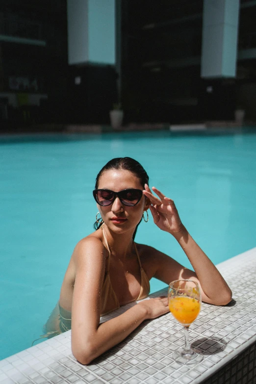 a woman sitting at the edge of a swimming pool, a portrait, pexels contest winner, holding a martini, orange sunglasses, instagram post, latinas