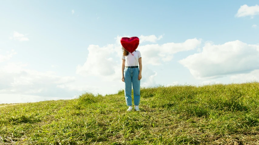 a woman standing on top of a lush green hillside, an album cover, inspired by Storm Thorgerson, pexels contest winner, magic realism, glass sculpture of a heart, inflatable, sadie sink, on a sunny day