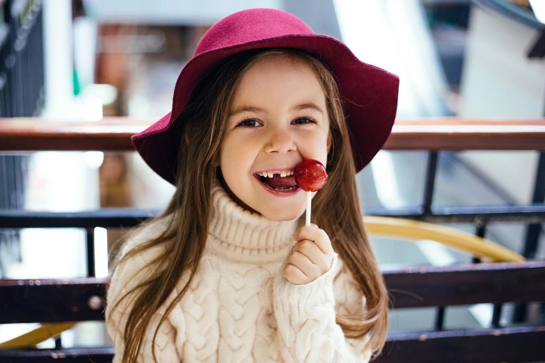 a little girl sitting on a bench eating a strawberry, by Elaine Hamilton, pexels contest winner, maroon hat, smiling fashion model, lollipop, wearing a red turtleneck sweater