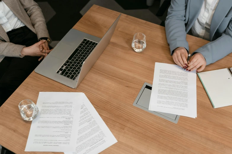 a couple of people sitting at a table with papers, on a wooden desk