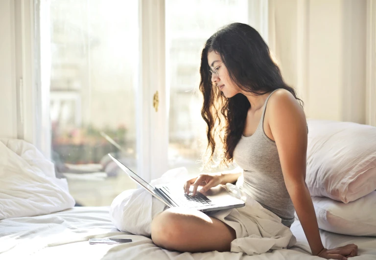 a woman sitting on a bed using a laptop computer, pexels, wearing a tanktop and skirt, wearing white pajamas, handsome girl, fine texutre