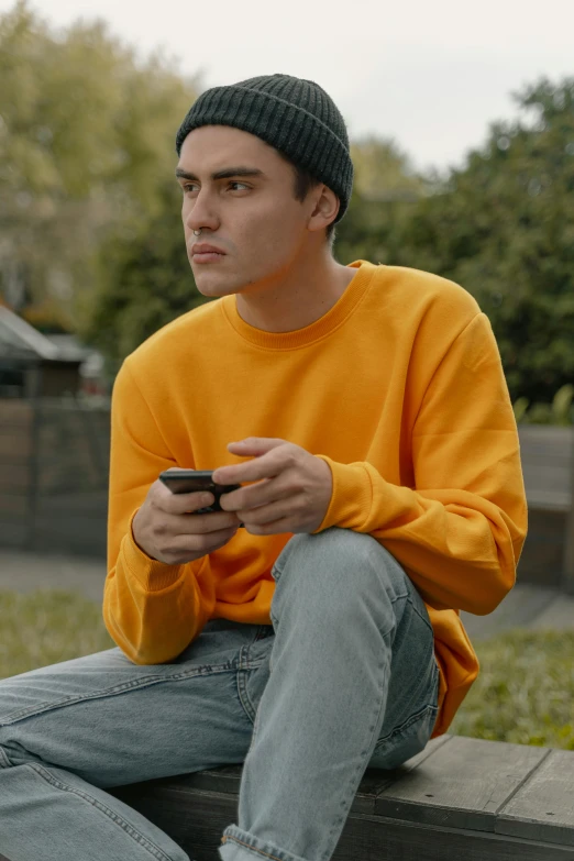 a man sitting on a bench looking at his cell phone, trending on pexels, long orange sweatshirt, yellow hue, federation clothing, awkward