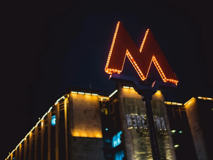 a large neon sign on the side of a building, by Adam Marczyński, pexels contest winner, modernism, black gotic letter m, moscow metro, 🦩🪐🐞👩🏻🦳, scenic view at night