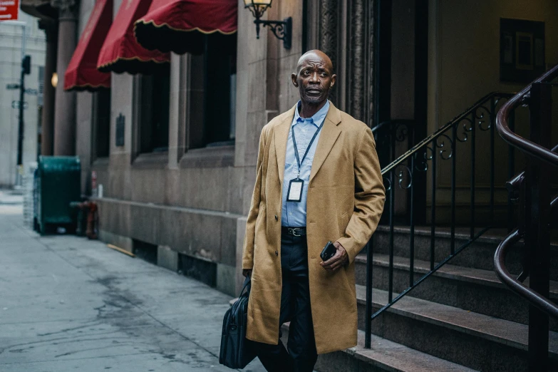 a man in a tan coat standing in front of a building, inspired by Henry O. Tanner, pexels contest winner, lance reddick, streets of new york, principal photography, full-body-shot