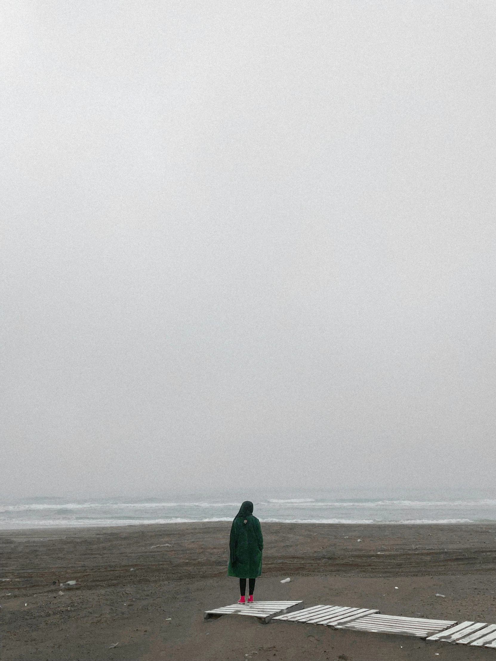 a person standing on a beach flying a kite, an album cover, by Caro Niederer, unsplash contest winner, postminimalism, dark green cloak, foggy rainy day, looking sad, wearing only a green robe