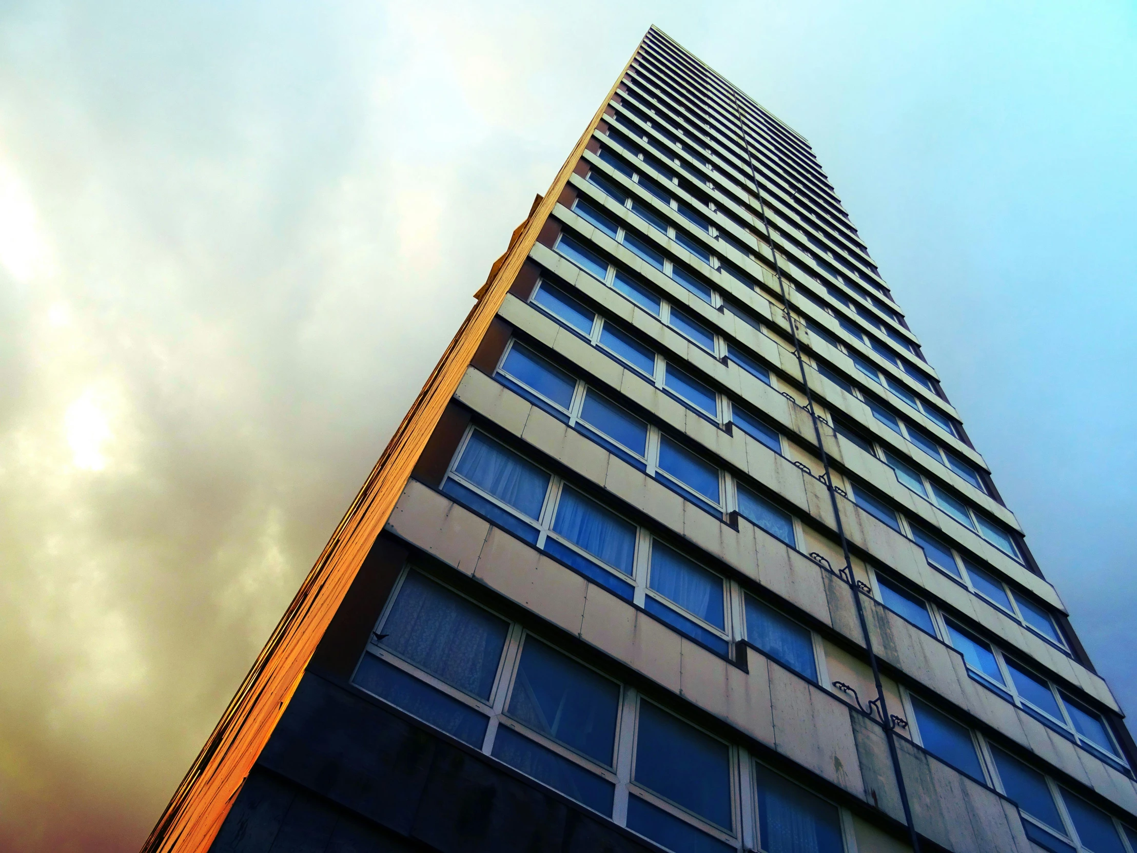 a tall building in front of a cloudy sky, an album cover, inspired by Richard Wilson, unsplash, brutalism, orange and blue sky, dramatic lighting - n 9, ten flats, view up