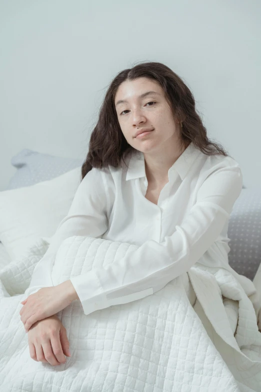 a woman sitting on top of a bed covered in a blanket, white dress shirt, louise zhang, no makeup wavy hair, wearing a white folkdrakt dress