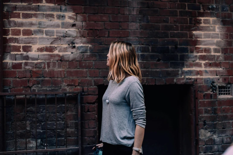 a woman standing in front of a brick building, inspired by Sydney Carline, minimalism, grey sweater, candid, dark backround, late morning