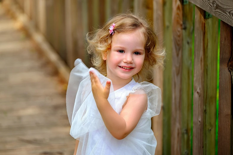 a little girl standing next to a wooden fence, inspired by Sophie Anderson, pixabay contest winner, wearing a white flowing dress, playful smile, portrait photography 4 k, pointing at the camera