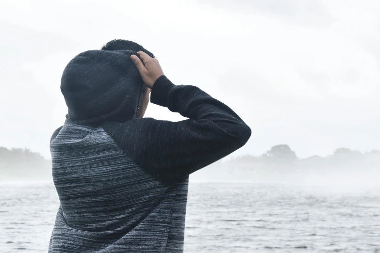a person standing in front of a body of water, grey hoodie, background image, pondering, overcast weather