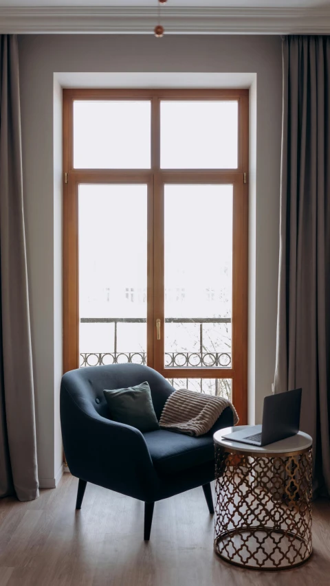 a chair sitting in a living room next to a window, inspired by Constantin Hansen, unsplash contest winner, paris hotel style, winter blue drapery, with a laptop on his lap, high arched ceiling