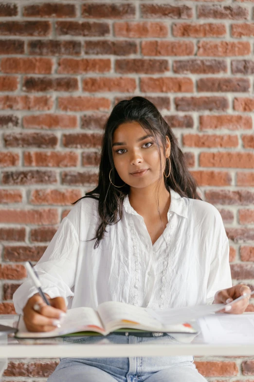 a woman sitting at a table with a book and pen, dressed in a white t shirt, indian girl with brown skin, in front of white back drop, wearing a white blouse