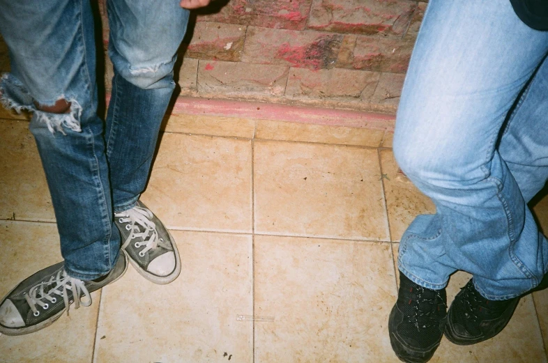 two people standing next to each other on a tiled floor, an album cover, inspired by Nan Goldin, unsplash, sots art, jeans and boots, many partygoers, exposed toes, very pale