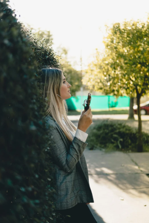 a woman standing on a sidewalk holding a cell phone, trending on unsplash, clad in vines, thoughtful pose, hedge, girl in suit