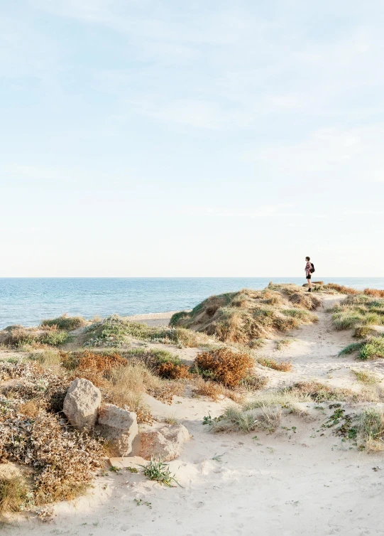 a man standing on top of a sandy beach next to the ocean, in a mediterranean lanscape, profile image, sports photo, girl walking between dunes