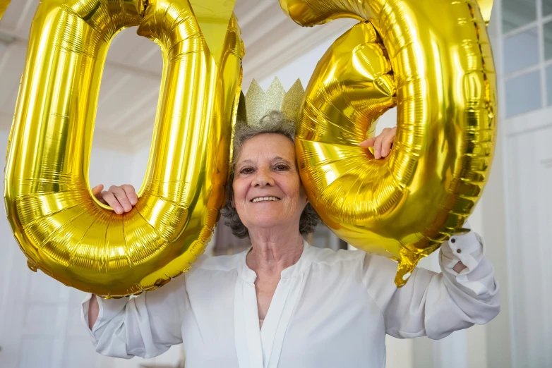 a woman holding two gold balloons in front of her face, pexels contest winner, 70 years old, sign that says 1 0 0, wearing a paper crown, avatar image