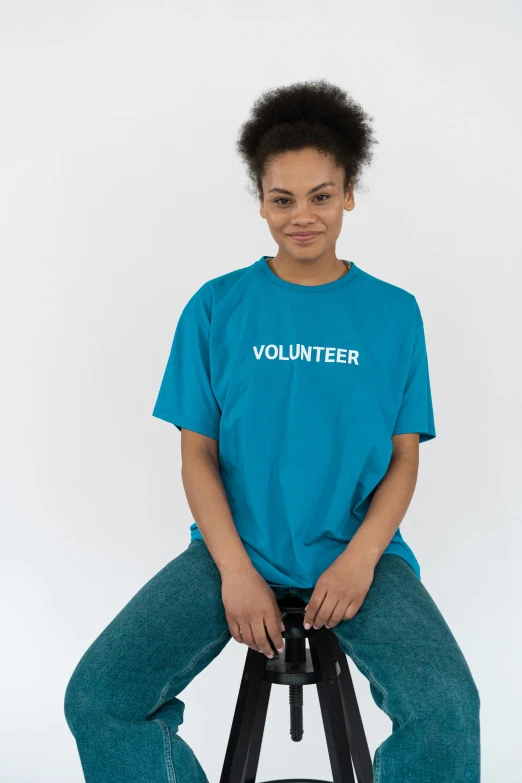 a woman sitting on a stool wearing a volunteer t - shirt, an album cover, trending on unsplash, teal uniform, product introduction photo, photographed for reuters, she is wearing streetwear