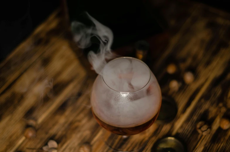 a glass filled with liquid sitting on top of a wooden table, trending on unsplash, renaissance, white thick smoke, cocktail in an engraved glass, thumbnail, from the elbow