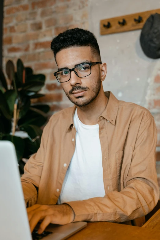 a man sitting in front of a laptop computer, by Cosmo Alexander, trending on pexels, renaissance, square rimmed glasses, androgynous male, middle eastern, wearing a brown