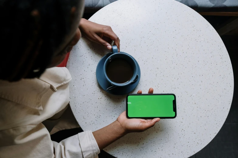 a woman sitting at a table holding a phone with a green screen, trending on pexels, coffee, ( ( dark skin ) ), square, 5k