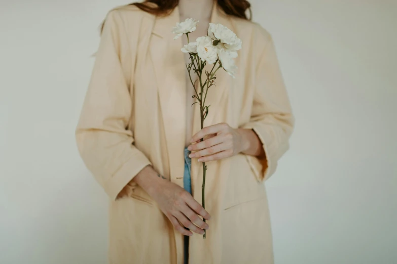 a woman holding a bunch of white flowers, trending on unsplash, aestheticism, wearing a long beige trench coat, wearing a light - pink suit, holding a yellow toothbrush, wearing silver silk robe